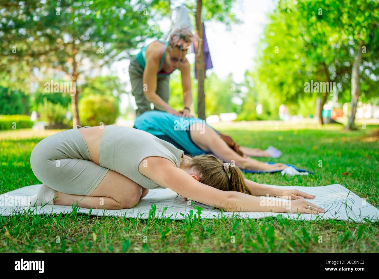 Le donne che indossano abbigliamento sportivo che si allungano e si rilassano nei bambini posano insieme sui tappeti sull'erba verde del parco. Bambini in posa, relax, yoga, erba, donne Foto Stock