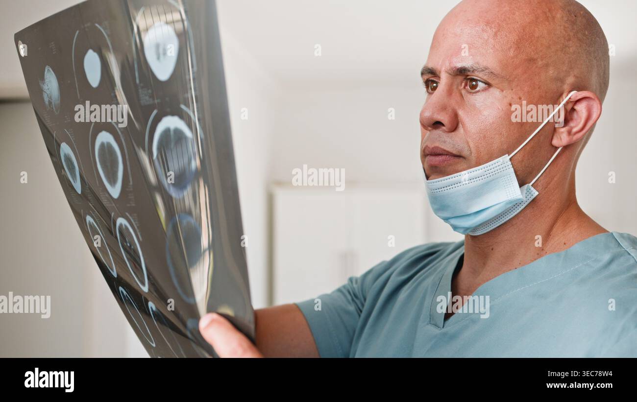 Medico maschio interessato che esamina la pellicola di scansione per MRI cerebrale in clinica con maschera chirurgica e scrub Foto Stock