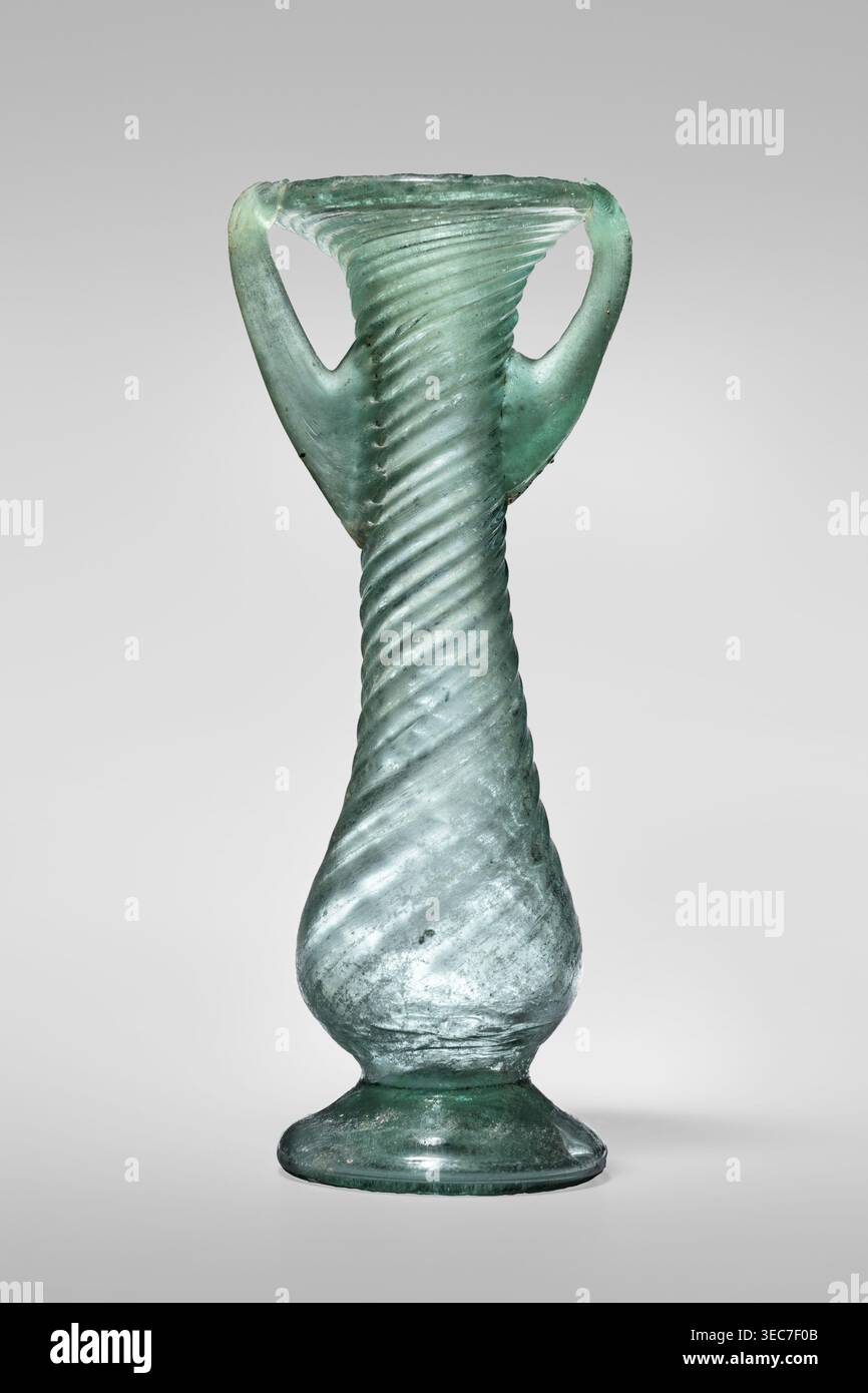 Unguentarium, sconosciuto laboratorio orientale. Recipiente in vetro, verde chiaro trasparente, Barag tipo VII: 1, 3. Unguentarium con pancia e collo larghi a forma di pera decorati con costole diagonali su tutta la superficie; piede con fessura; due morse verticali attaccate a due terzi dell'altezza del recipiente e che si estendono fino al bordo del bordo a forma di imbuto. Libero. Diametro esterno del cerchione 4,50 cm; altezza 12,50 cm; diametro esterno inferiore 4,30 cm; peso 0,072 kg. III-IV secolo, Siria., Un vaso di vetro traslucido con una struttura a spirale attorcigliata che corre verticalmente lungo il suo corpo, affusolato verso un collo più stretto e. Foto Stock