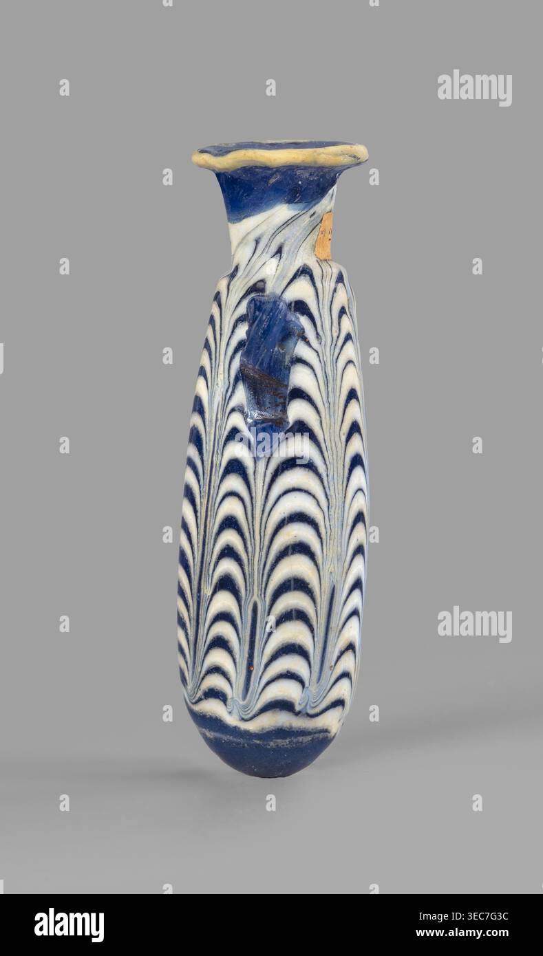 Alabastron, laboratorio egiziano, vi-V secolo a.C. Vetro blu scuro opaco decorato con fili a zigzag gialli-marroni; corpo ovoide, fondo arrotondato, bordo discoidale piatto, morse d'anatra verticali incollate alla parte superiore del corpo; formate sul nucleo. Tipo: Grose Group i:e, Form i:6, Harden Group i Form 4. Vaso di Alabastron, altezza 8,6 cm; diametro del corpo 2,5 cm; diametro del beccuccio 2,45 cm; peso 0,030 kg; luogo di origine egiziano?.. Un vaso di vetro stretto e allungato con bordo svasato e base arrotondata su uno sfondo grigio neutro, decorato con un modello simile a piume ripetuto di profondità alternata Foto Stock