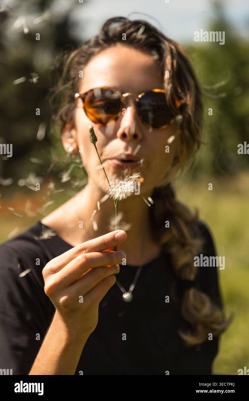 Una giovane donna si trova in un campo verde con gli occhiali da sole. Tiene in mano un dente di leone e soffia dolcemente, inviando semi nell'aria. Il sole splende brillantemente Foto Stock