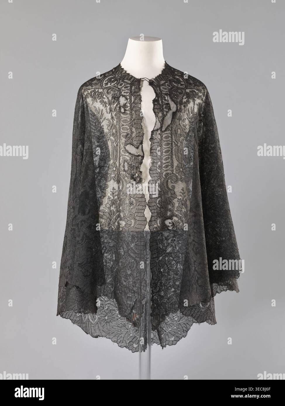 Capo - mantilla; scialle; provenienza dell'acquisto; moda; abbigliamento femminile; accessorio di abbigliamento; mantello in pizzo mantilla, triangolare. Schema: Ghirlande di foglie sul fondo, alberi conici nelle curve sul fondo. Alla fine del XIX secolo, in Francia. Pizzo; dimensioni: Lunghezza 96 cm, larghezza 344 cm., mantello in pizzo nero trasparente visualizzato su un busto bianco di manichino su sfondo grigio neutro, con motivi floreali e a spirale ornati, bordi smerlati lungo l'orlo e l'apertura frontale, un'alta scollatura arrotondata e un'apertura frontale centrale con delicati rifiniture arricciate; il pizzo mostra diverse densità Foto Stock