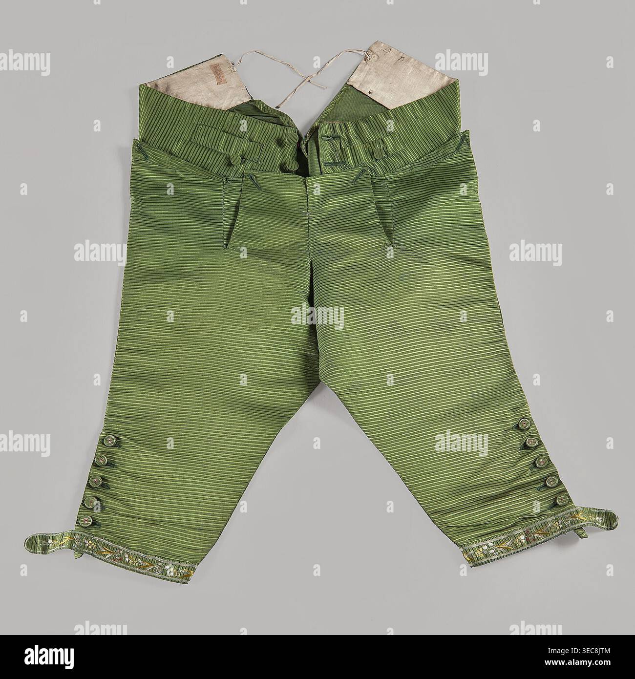 Pantaloni corti al ginocchio con patta, realizzati in tessuto di seta verde. Due piccole tasche sotto la patta, grandi tasche sui lati e due piccole tasche per portachiavi sopra la patta. Estremità gambe con polsini stretti fissati ai lati con sei bottoni ricamati. Schienale regolabile, legato con un nastro. Motivi vegetali e floreali. xviii secolo, anni '1770 Slesia; prodotto in Francia. Abbigliamento da uomo; lunghezza 81 cm., Un paio di pantaloni corti realizzati in tessuto a righe verdi appoggiati su uno sfondo grigio, con vita alta con fodera interna bianca e corde legate, linguette anteriori in vita con tessuto- Foto Stock