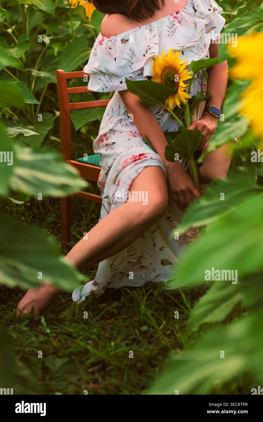 Figura femminile che regge un girasole seduto nella natura circondato dal verde e dalla luce soffusa. Il momento sereno mette in risalto la consapevolezza del corpo, il sé Foto Stock