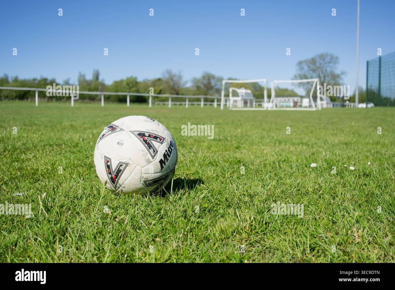 Aprile 2026, Northamptonshire, Regno Unito: Un basso punto di vista di un calcio sull'erba. In lontananza, ci sono bocche e ringhiere intorno a un campo da calcio. Foto Stock