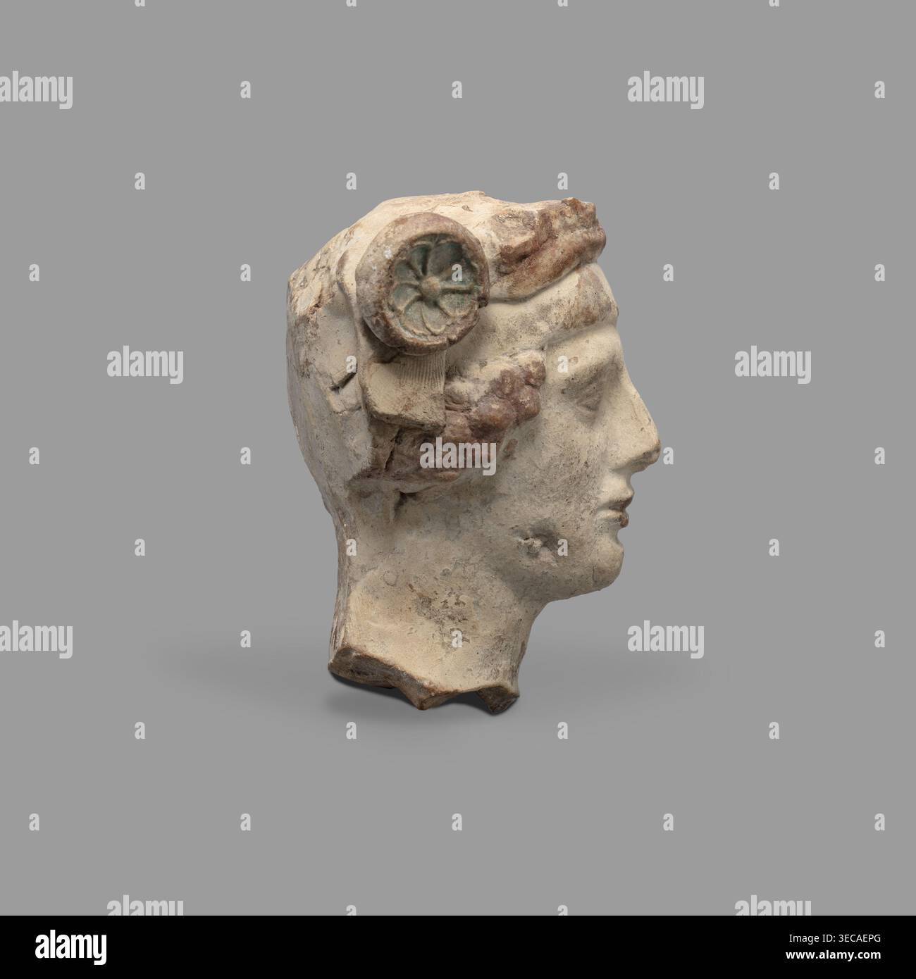 Capo di una statuetta di Dioniso. Testa di un giovane uomo con un volto privo di espressione, con una bocca ampia e compatta, un volto triangolare con guance intagliate, occhi grandi, naso prominente e sopracciglia delicatamente delineate. Sulla sua testa il giovane indossa un diadema decorato con rosette ai lati e una fitta corona. Tracce di vernice rossa (cinabro) su capelli, labbra e diadema. Blu sulle rosette — negli incavi. V-IV secolo a.C., Taranto, Italia meridionale (Italia). Statuetta in terracotta, altezza 8,7 cm, larghezza 8,3 cm, profondità 6,3 cm, peso 0,28 kg. Autore: Scen. Galloni Classe; iscrizione/ Foto Stock