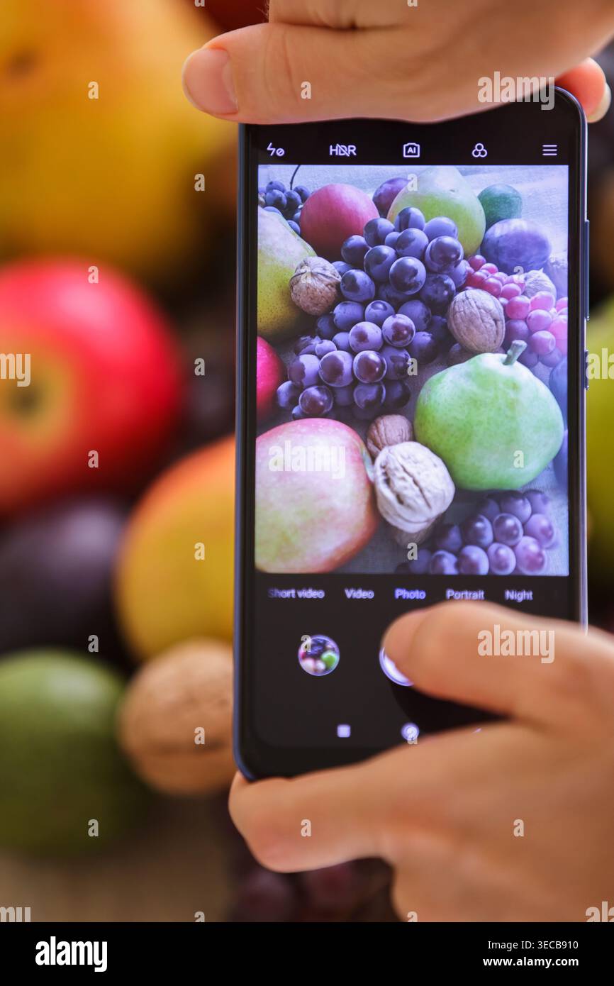 Primo piano verticale dello schermo di uno smartphone che cattura la natura morta di uva, pere e noci. Le mani di una persona sono visibili tenendo di nuovo il dispositivo Foto Stock