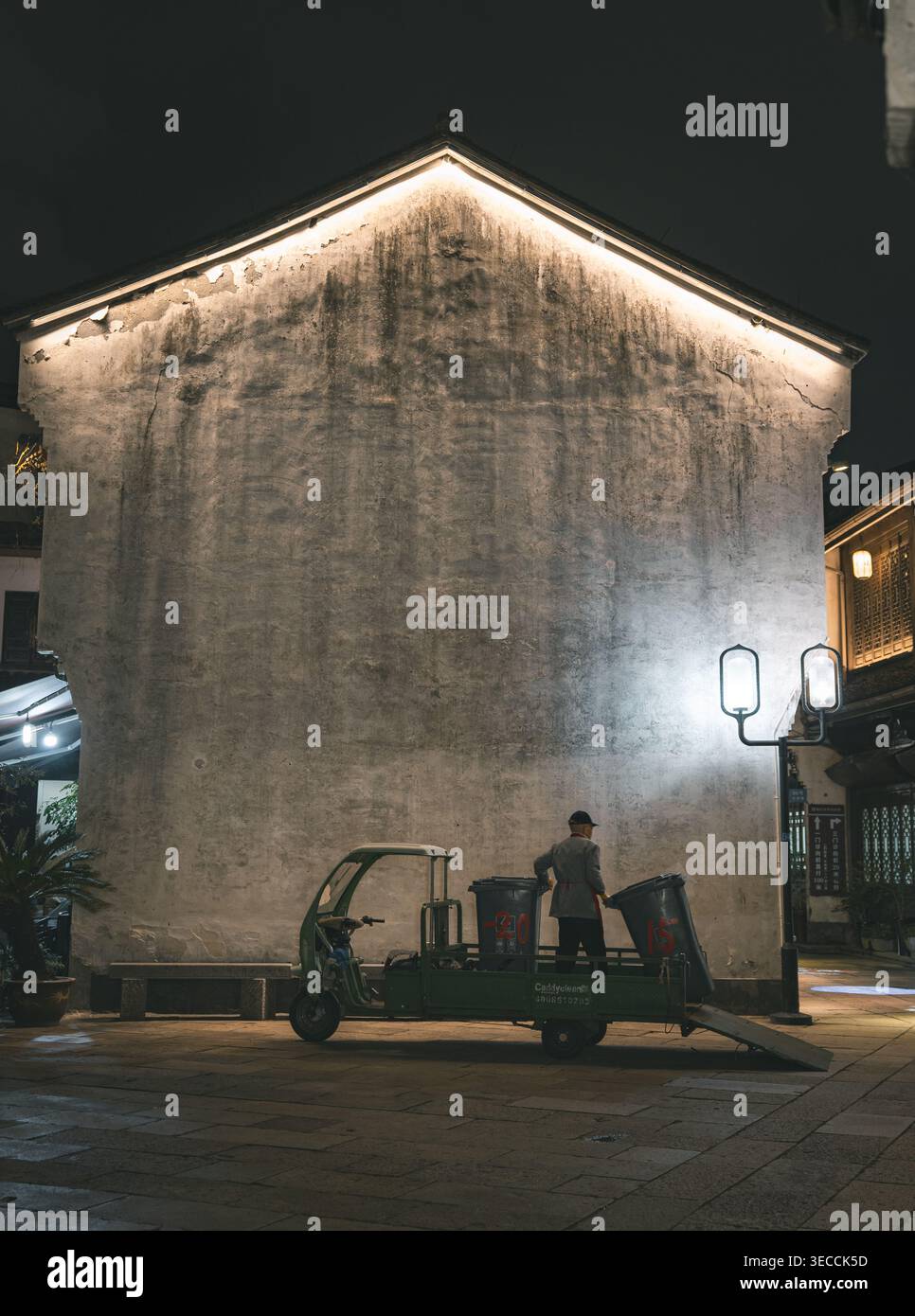 Hangzhou, Cina - 11 novembre 2025 Un lavoratore si trova a fianco di un veicolo multiuso verde a tre ruote con cassonetti, illuminato da lampioni e edifici Foto Stock