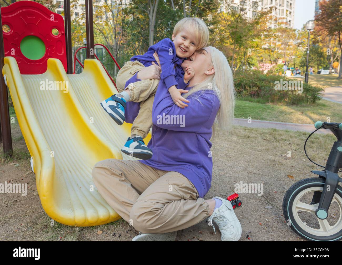 Madre e bambino giocano insieme in un parco in una giornata di sole vicino a un parco giochi mentre si divertono sullo scivolo Foto Stock