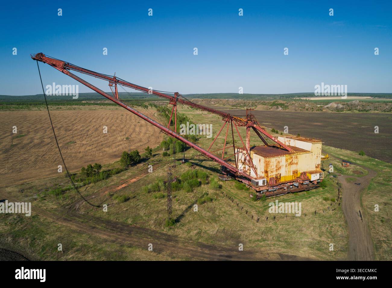 L'enorme escavatore dragline industriale poggia in un campo aperto, circondato da vegetazione sparsa e strade sterrate Foto Stock