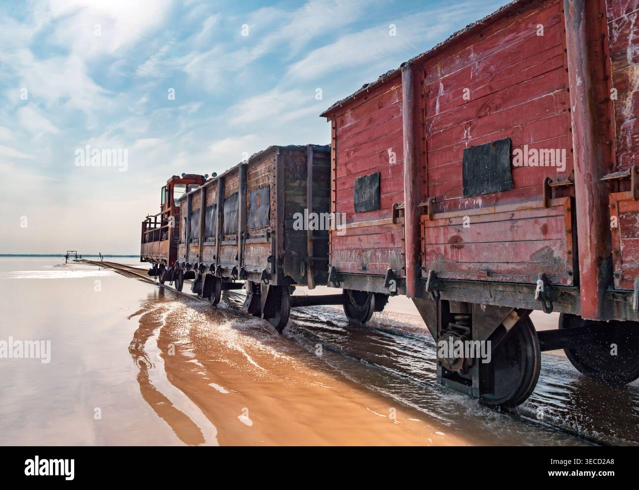 Locomotiva industriale Rusty che traina carri merci in legno attraverso acque rosa poco profonde a Bursol Lake Foto Stock