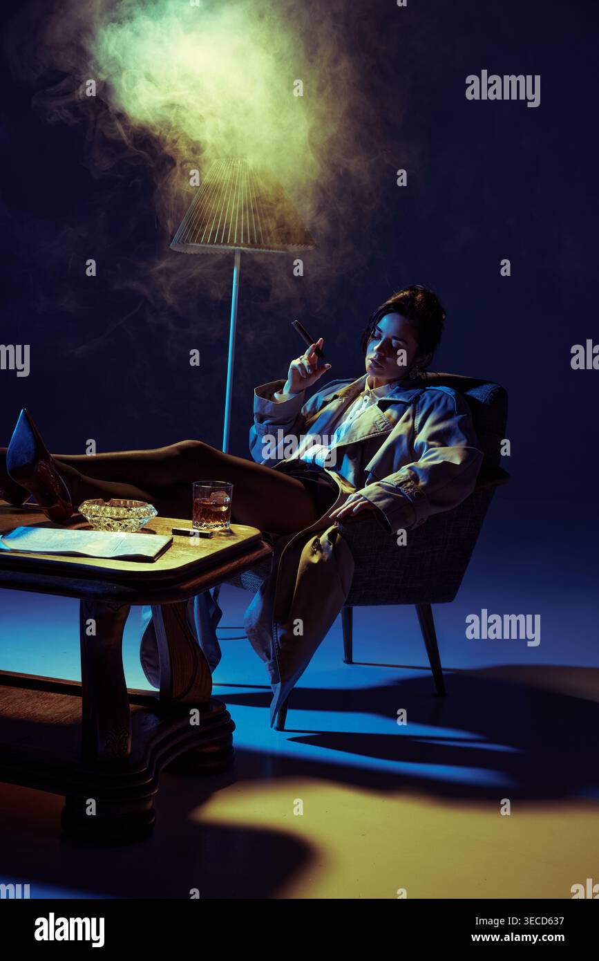 Donna che fuma sigaro e si rilassa con il whisky in poltrona sotto l'illuminazione colorata dello studio. Foto Stock