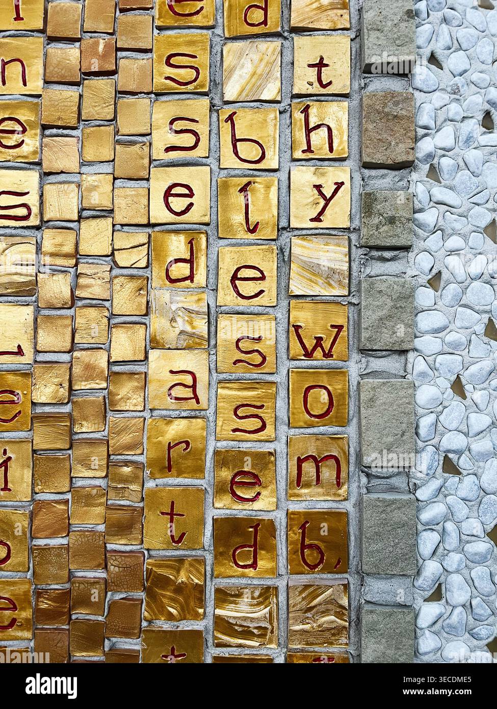 Piastrelle decorative in oro a mosaico con lettere dipinte di rosso e bordo in pietra di ciottoli che creano un'arte tipografica testurizzata delle pareti. Foto Stock