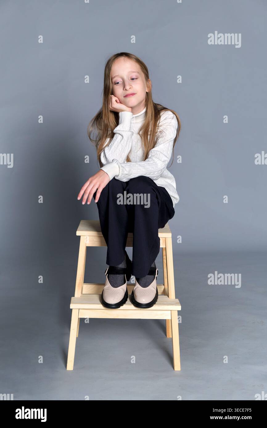 La ragazza siede sullo sgabello in legno con le mani delicatamente appoggiate sul ginocchio, un sorriso sottile e una tranquillità Foto Stock