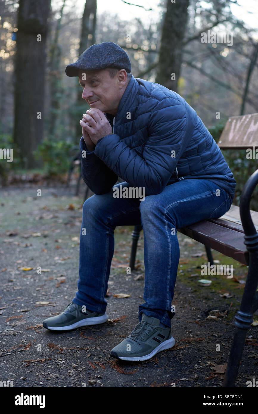 Uomo di mezza età seduto su una panchina del parco inclinato in avanti con le mani strette e un sorriso sottile, guardando lateralmente in un umore premuroso e fiducioso Foto Stock