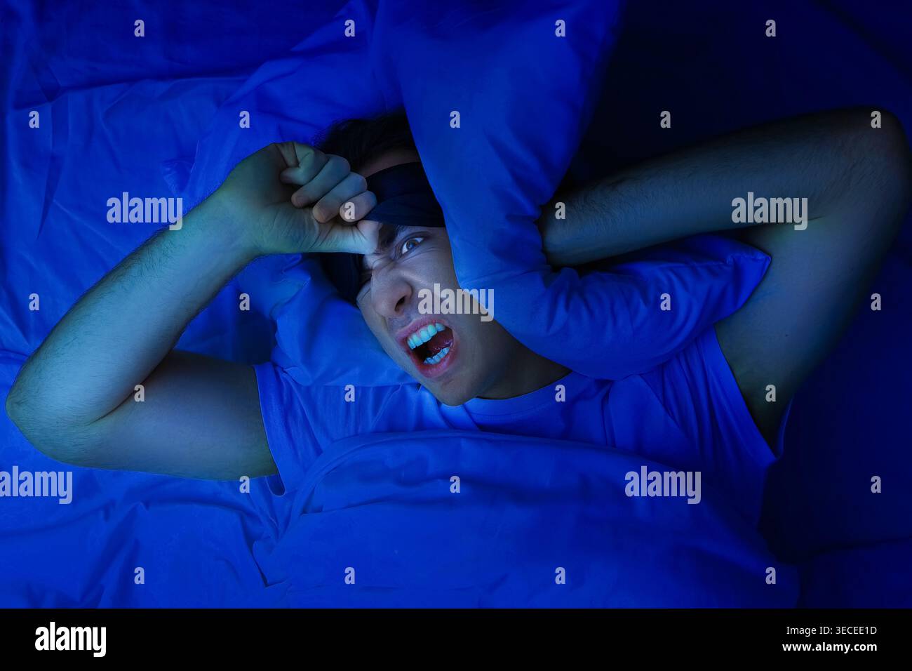 Un uomo arrabbiato con una maschera dormiente copre le orecchie con un cuscino, soffre di insonnia, rumore e privazione del sonno in una camera blu scuro di notte. H Foto Stock