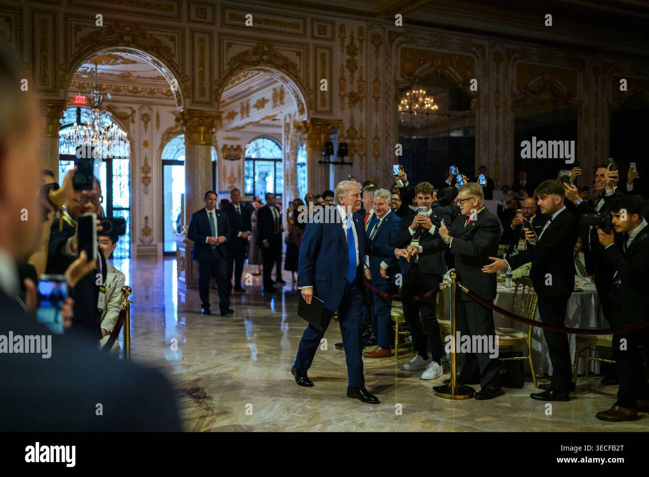 Il presidente Donald J. Trump saluta i partecipanti mentre cammina attraverso una sala interna ornata presso il Mar-a-Lago Club prima di parlare a una conferenza sulla criptovaluta a Palm Beach, Florida, il 25 aprile 2026. Per gentile concessione della Casa Bianca. Foto Stock