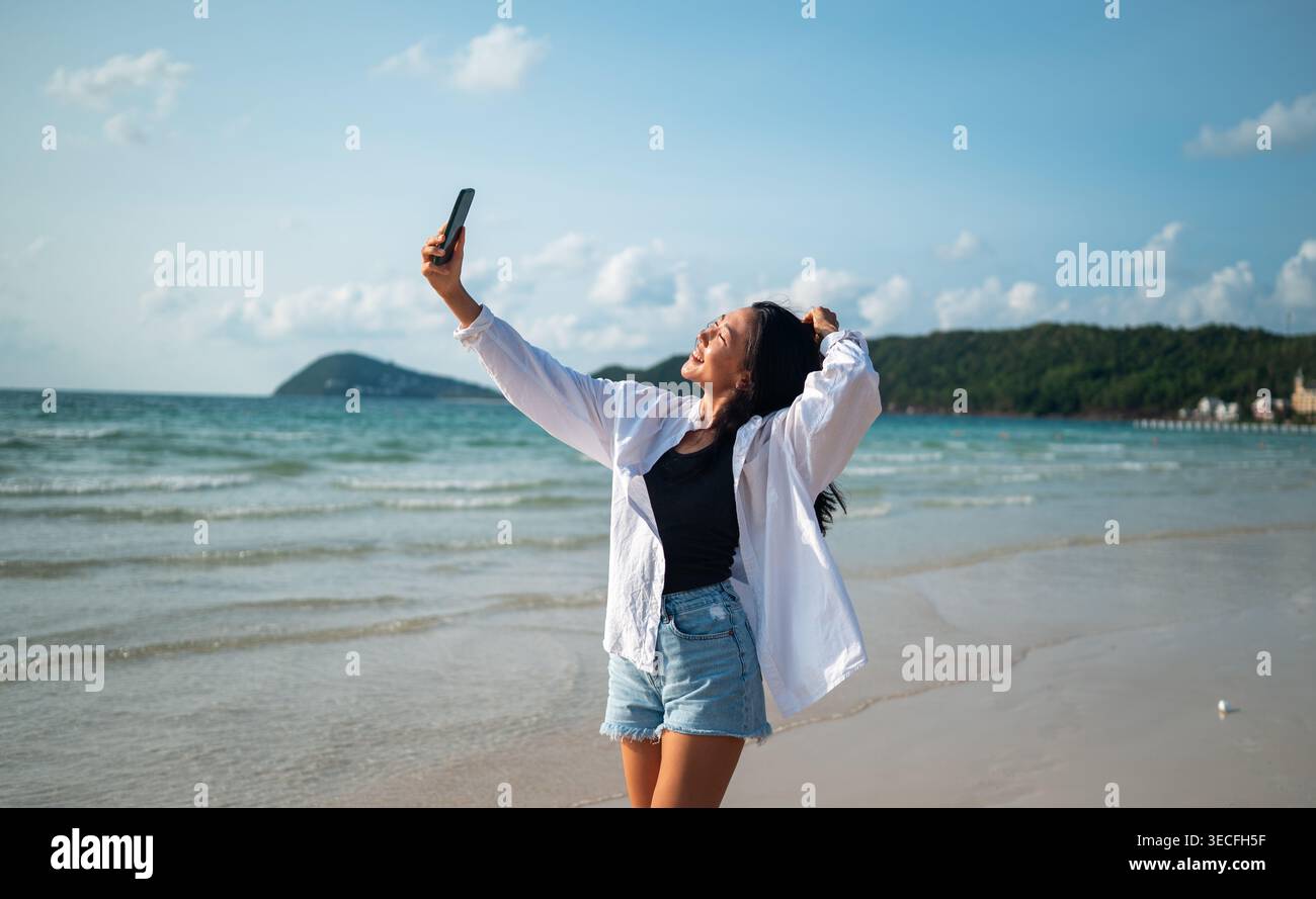 Influencer che scatta un selfie su una spiaggia tropicale, si gode una vacanza, crea contenuti sui social media Foto Stock