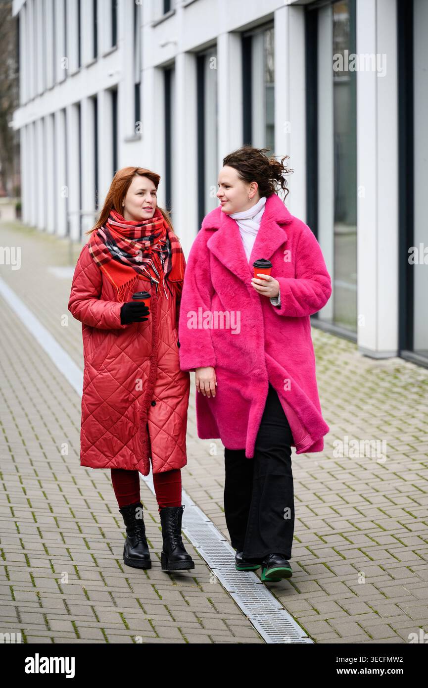 Due sorelle felici che camminano per la strada della città e bevono un caffè. Giovani donne eleganti con cappotti rosa e rosso brillanti che parlano mentre passeggiano all'aperto. Conce Foto Stock