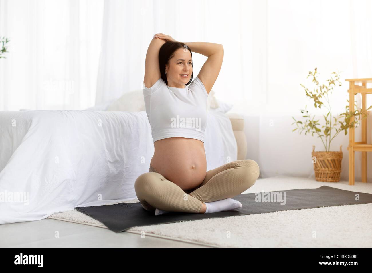 Donna attiva in gravidanza seduta su un tappetino da yoga che si allunga le braccia sopra la testa con un sorriso Foto Stock