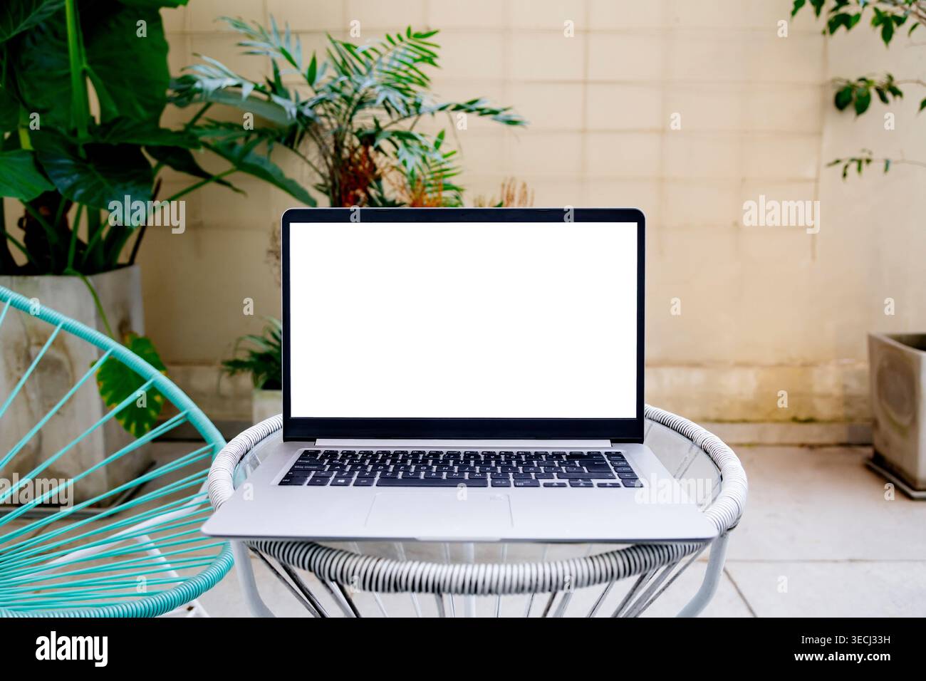 Computer portatile per il trucco con schermo bianco vuoto sul tavolo esterno nel moderno patio giardino con piante tropicali e sedia Acapulco Foto Stock