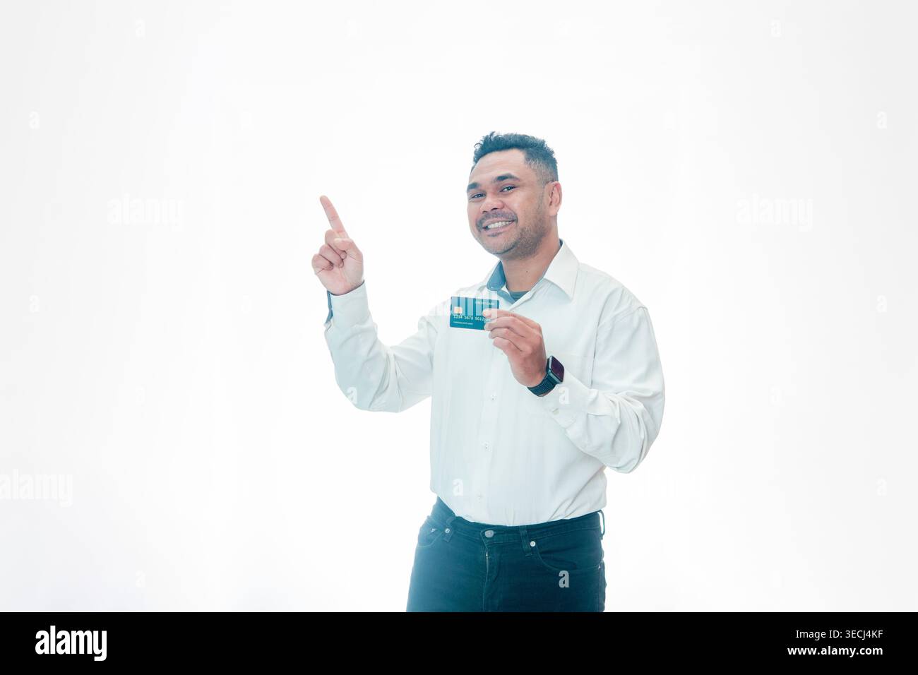 Un uomo indonesiano sorridente in camicia bianca che tiene una carta di credito e indica verso l'alto, presentando una soluzione finanziaria o un servizio bancario, isolato su bianco. Foto Stock