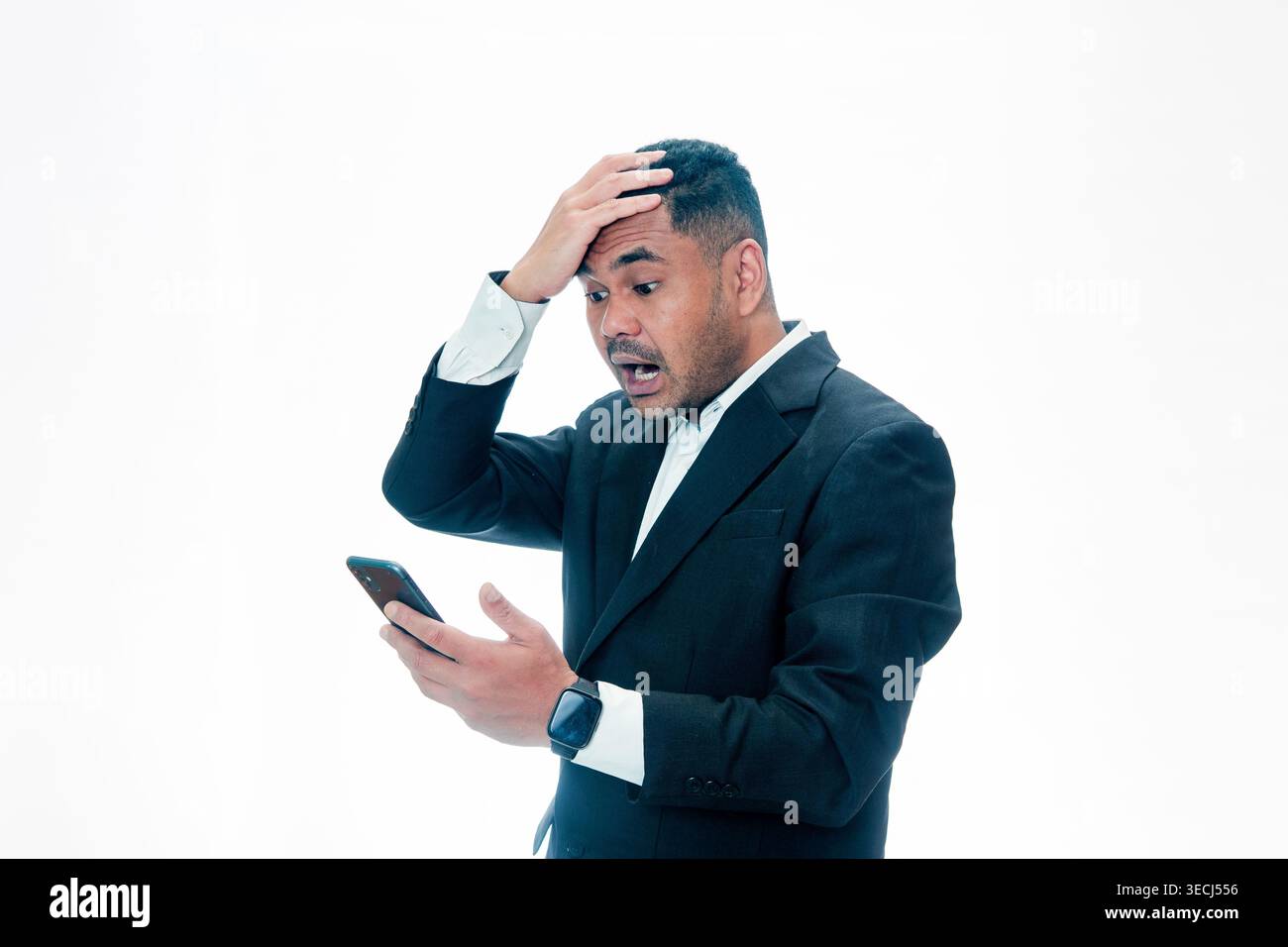 Un uomo indonesiano in abito nero che guarda il suo smartphone con un'espressione scioccata e si batte la testa con una mano, isolato su uno sfondo bianco. Foto Stock