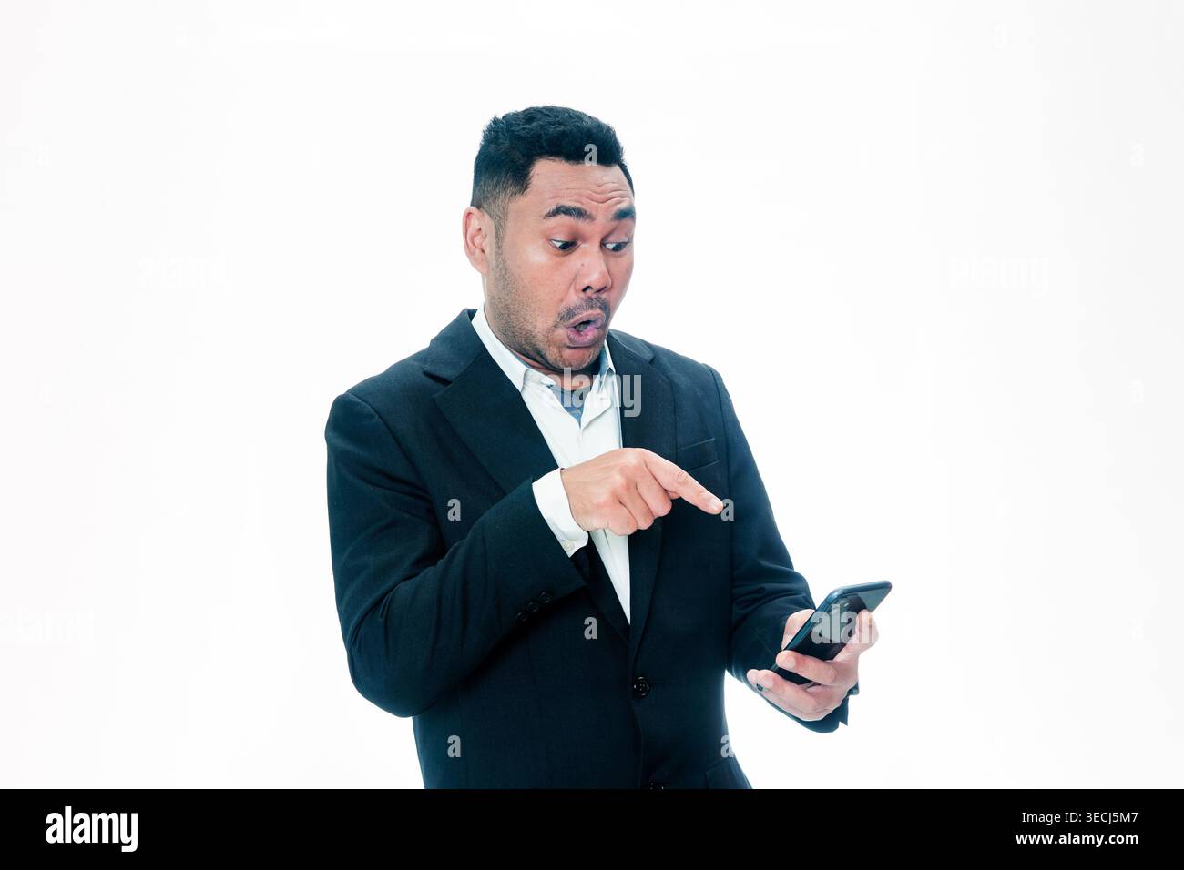 Un uomo indonesiano in abito nero che guarda il telefono con un'espressione scioccata e indica lo schermo con il dito su uno sfondo bianco. Foto Stock