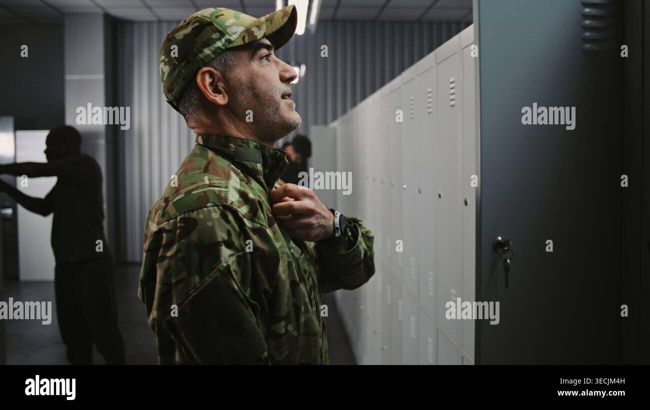 Il soldato maschile con braccia tatuate indossa la giacca e il berretto in uniforme OCP mimetico di Locker, preparandosi per la missione tattica mentre i colleghi interagiscono in background. Preparazione della base militare. Foto Stock