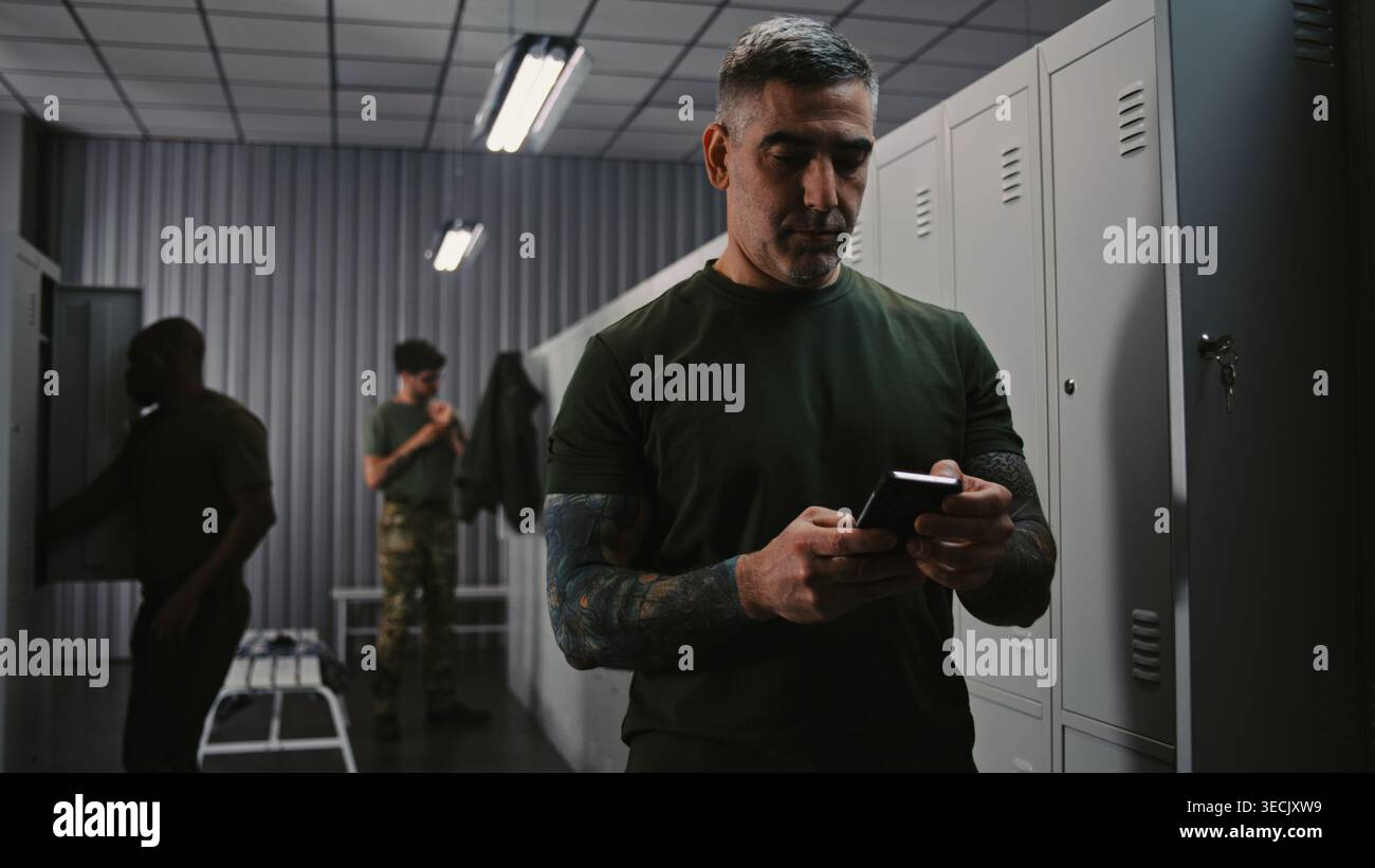 Athletic Military Man in Camouflage Cap and Tactical Gear utilizza lo smartphone per inviare aggiornamenti importanti o contattare i parenti presso l'area degli armadietti della base dell'esercito. Concetto di Modern Warfare Communication. Foto Stock