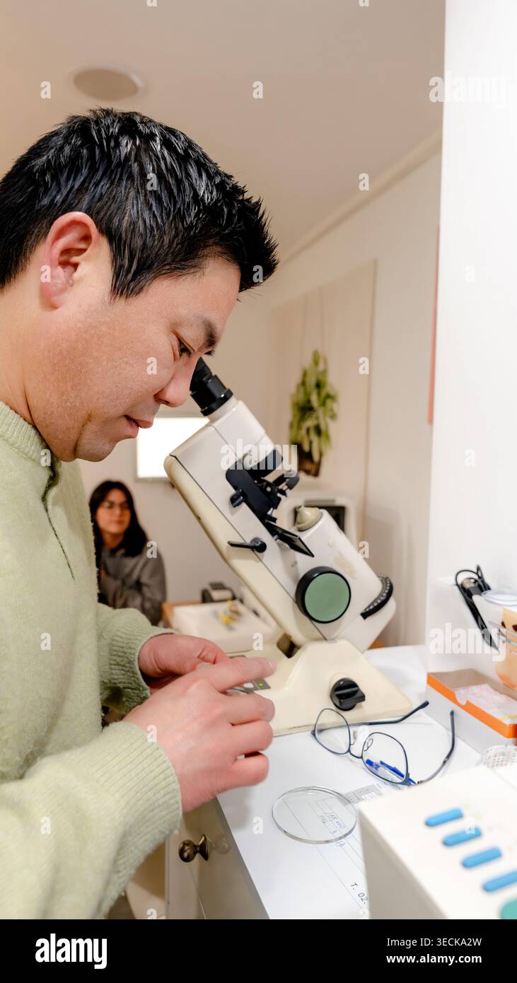 Un uomo esamina attentamente una lente ottica presso una stazione per microscopi in un centro ottico, mostrando precisione e dedizione per garantire la soddisfazione del cliente Foto Stock