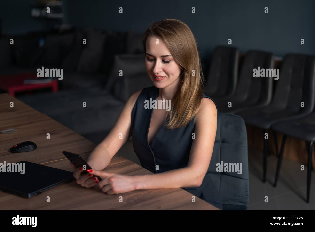 Giovane donna d'affari seduta a una scrivania moderna, sorridente mentre utilizza uno smartphone accanto a un laptop. Ambiente di lavoro professionale, produttività, Foto Stock