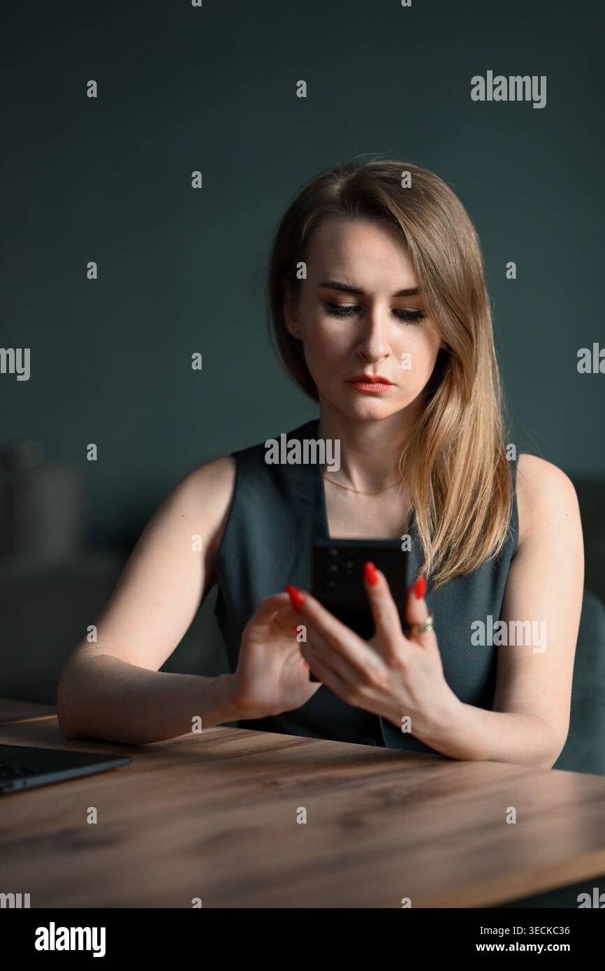 Donna d'affari concentrata che lavora a una scrivania in un ufficio moderno, controlla i messaggi su uno smartphone accanto a un laptop, attenta e professionale a bassa pressione Foto Stock