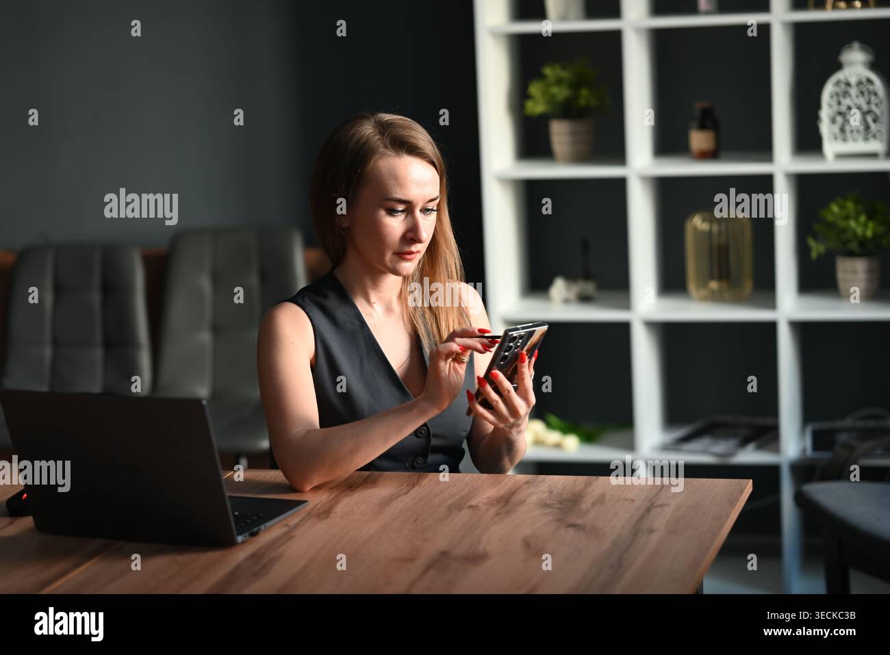 Donna d'affari concentrata su una scrivania di legno che utilizza uno smartphone accanto a un computer portatile, lavora in un ufficio moderno con scaffali, piante e luce soffusa. Foto Stock