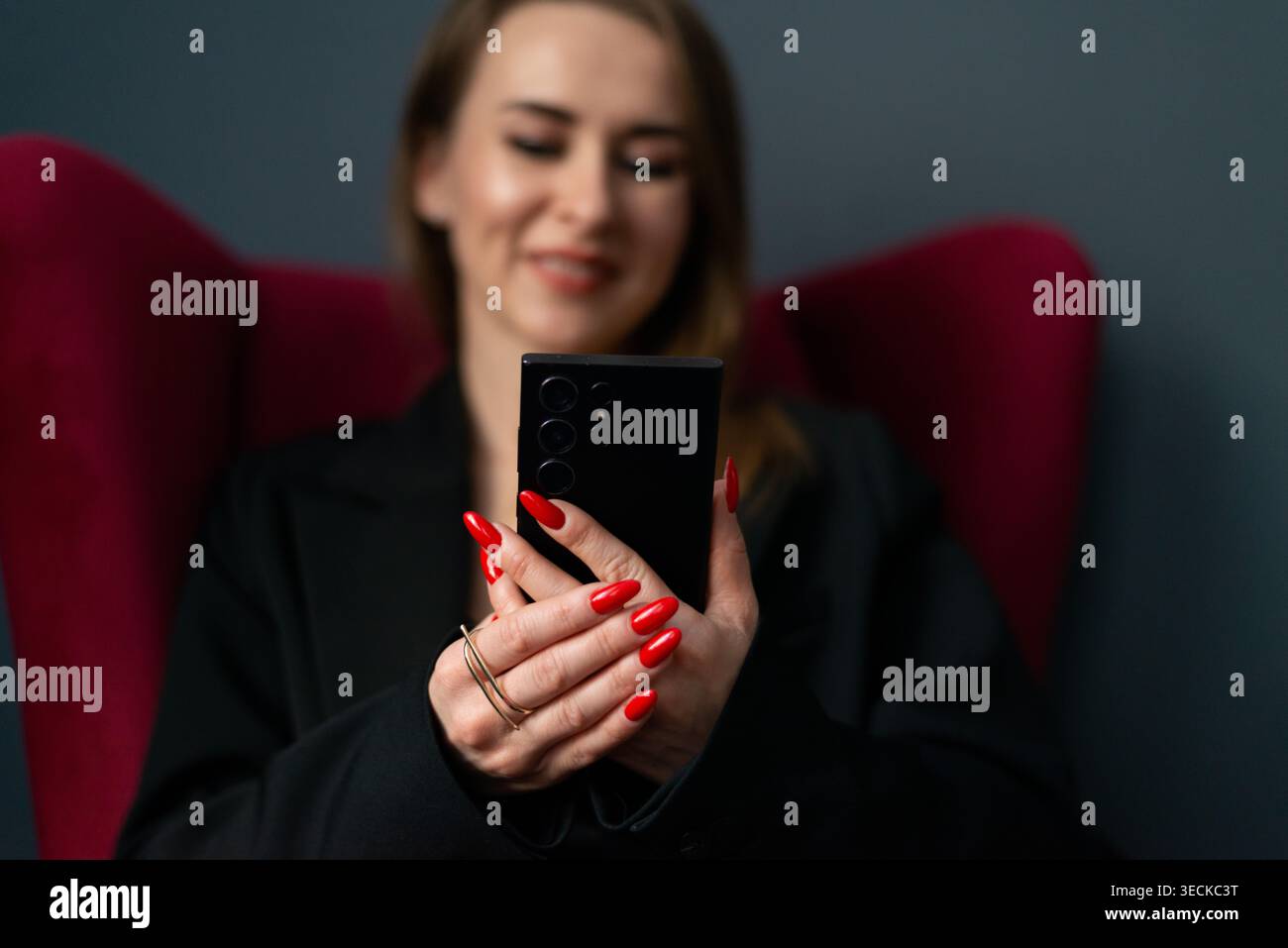 Donna d'affari sorridente in blazer che utilizza uno smartphone mentre si siede su una moderna sedia da ufficio, che mostra comunicazione mobile, lavoro remoto, produttività, Foto Stock
