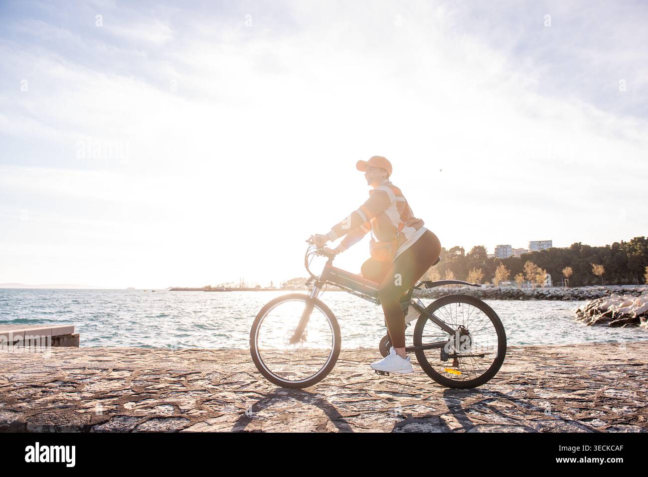Vista laterale di una donna che pedala su una bicicletta elettrica lungo una passeggiata sul lungomare all'alba, retroilluminata dal sole caldo, con mare, rocce e skyline della città Foto Stock