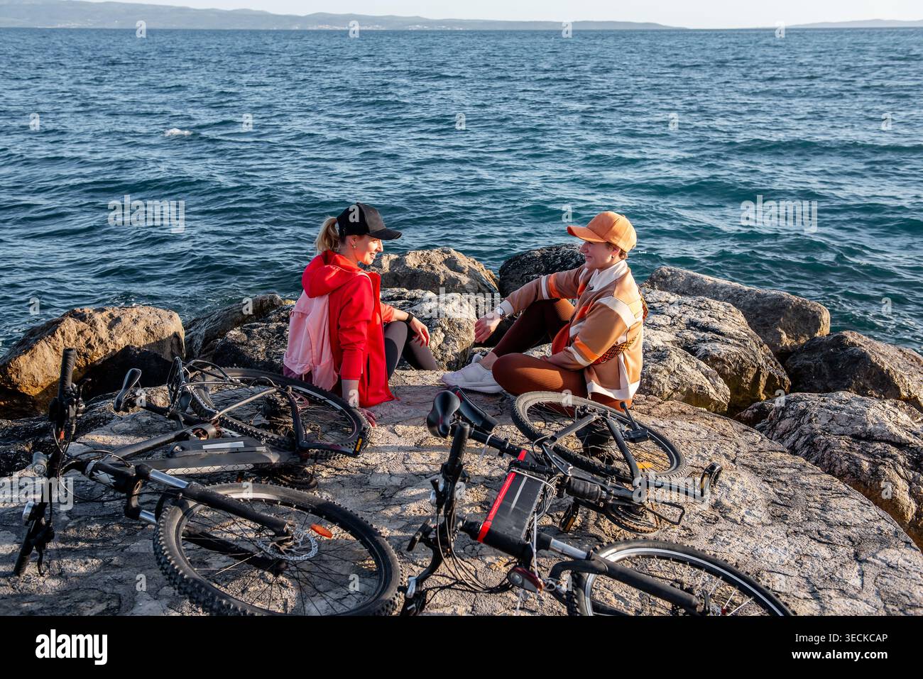 Due amiche si rilassano sulle rocce costiere accanto alle biciclette elettriche, chiacchierando dopo un giro sportivo all'aperto. La vista sull'oceano e l'aria fresca catturano un noi attivo Foto Stock