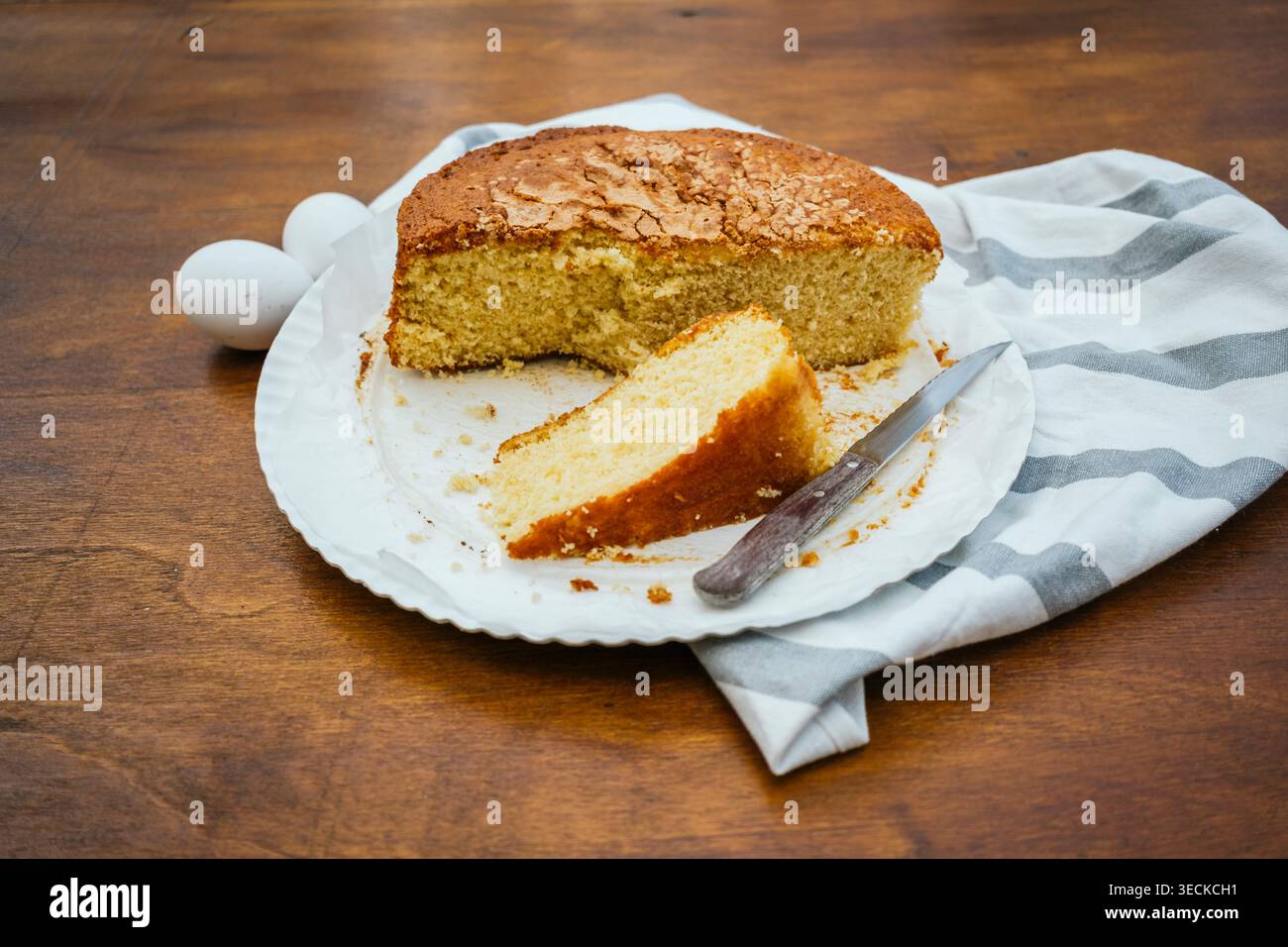 Torta rustica fatta in casa con un pezzo a fette su un piatto bianco, coltello e briciole accanto, sistemata su un tavolo di legno con un asciugamano da cucina a righe e un uovo Foto Stock