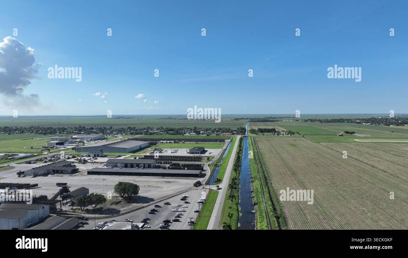 Vista aerea di grandi magazzini industriali e vasti campi agricoli divisi da un canale d'acqua dritto sotto un cielo blu con una grande nuvola bianca. Foto Stock