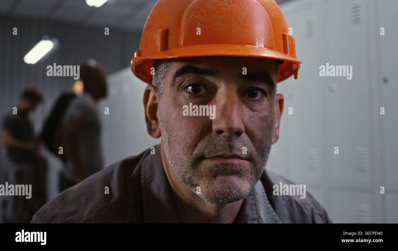 Primo piano di un esperto operaio in fabbrica con giacca da lavoro grigia e casco arancione, seduto sul banco e guardando il telaio con armadietti in metallo e colleghi in background. Preparazione industriale. Verticale. Foto Stock