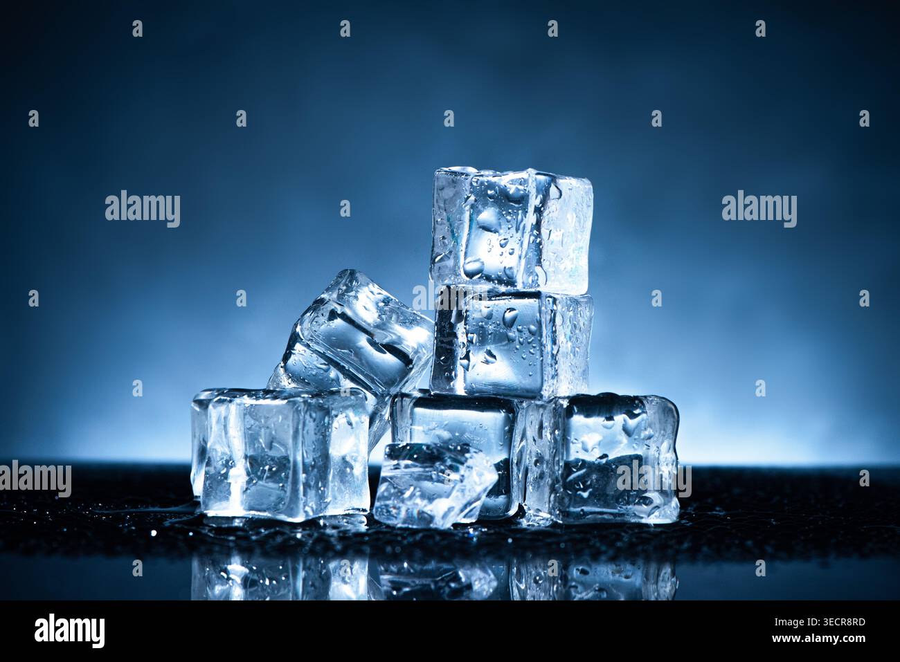 I cubetti di ghiaccio si fondono, mostrando gocce d'acqua su una superficie scura riflettente su uno sfondo blu Foto Stock