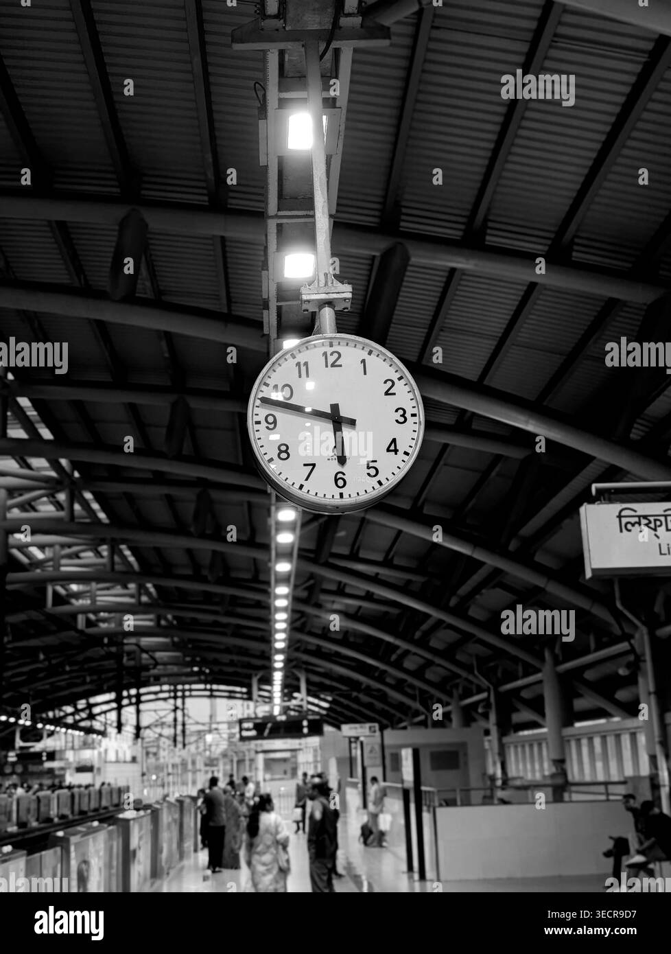 Vista in bianco e nero dell'interno di una stazione ferroviaria con un grande orologio sospeso, i passeggeri che aspettano sotto e le linee di luci principali che creano una profondità di - Immagine stock catturata con smartphone