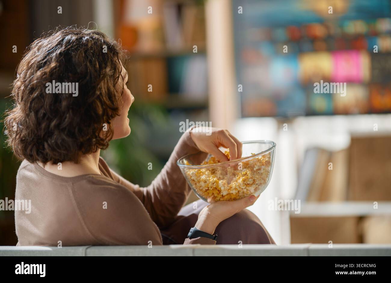 Ambiente di Home Entertainment rilassato con una donna che si gusta una ciotola di popcorn freschi mentre guarda la televisione. L'atmosfera e' intima e invitante, hi Foto Stock