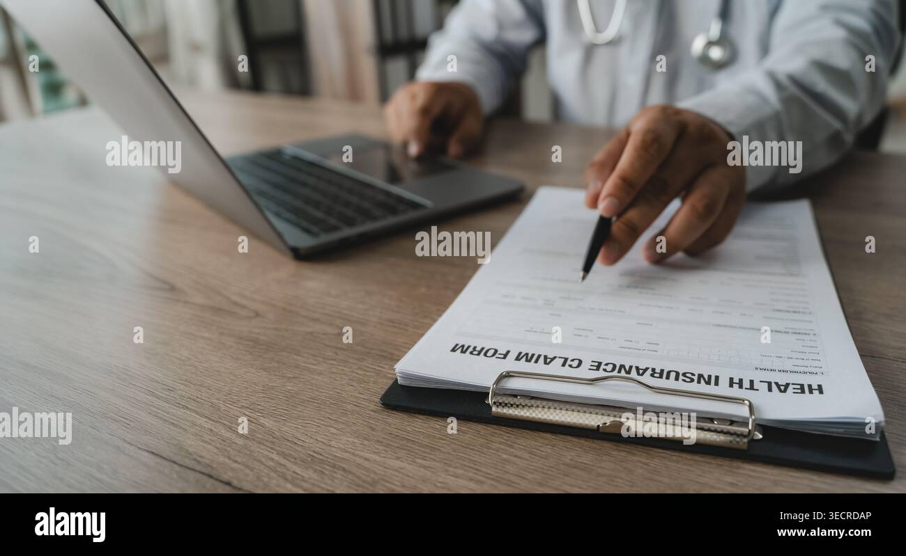 Mano che tiene la penna che esamina il documento di richiesta di assicurazione sanitaria accanto al laptop e allo stetoscopio che esegue la verifica dei documenti per i conti delle spese mediche Foto Stock