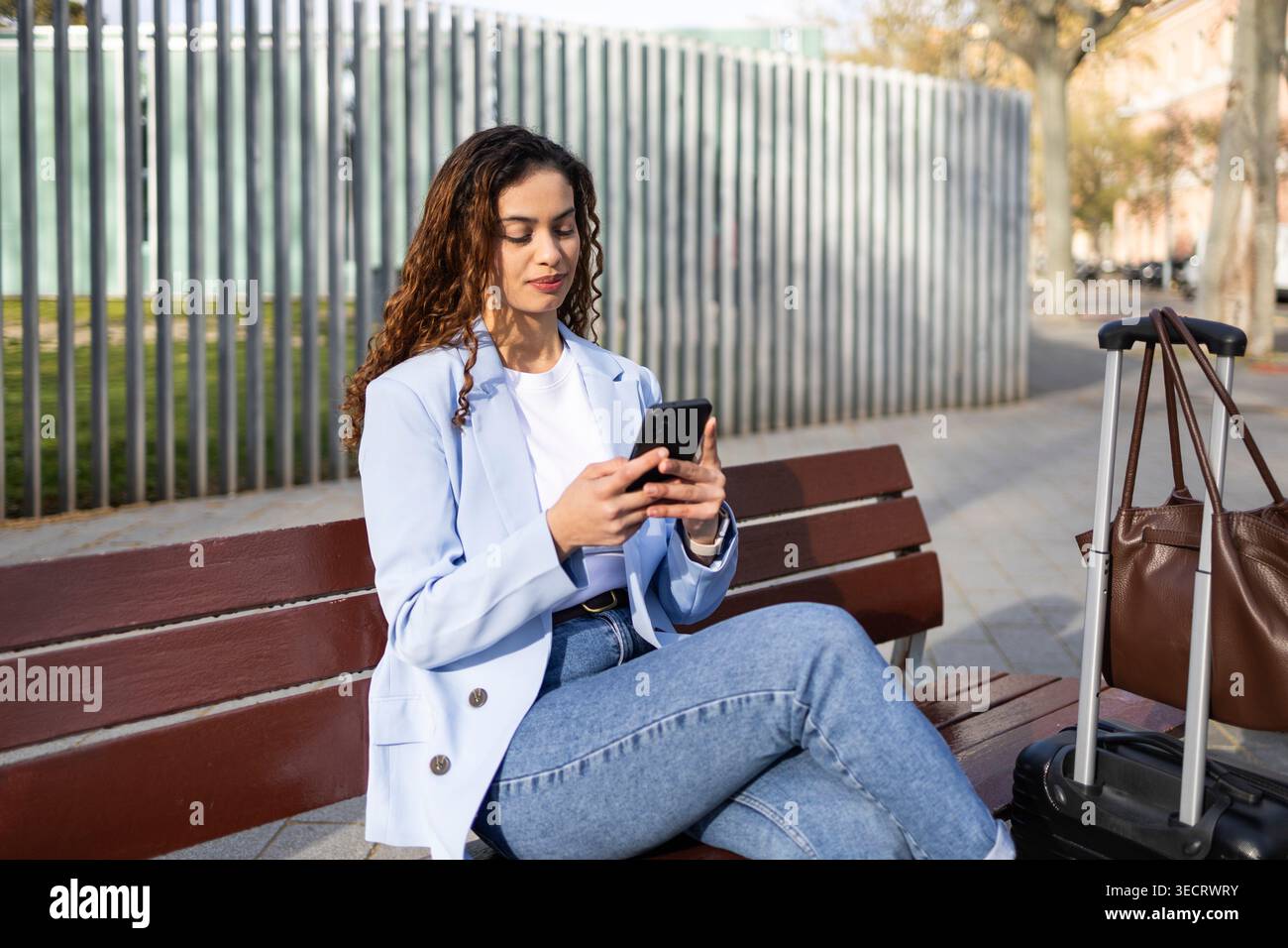 Donna d'affari seduta su una panchina con valigia e borsa, controllando il suo smartphone e i piani di viaggio all'aperto, preparandosi per un viaggio d'affari e per una coordina Foto Stock