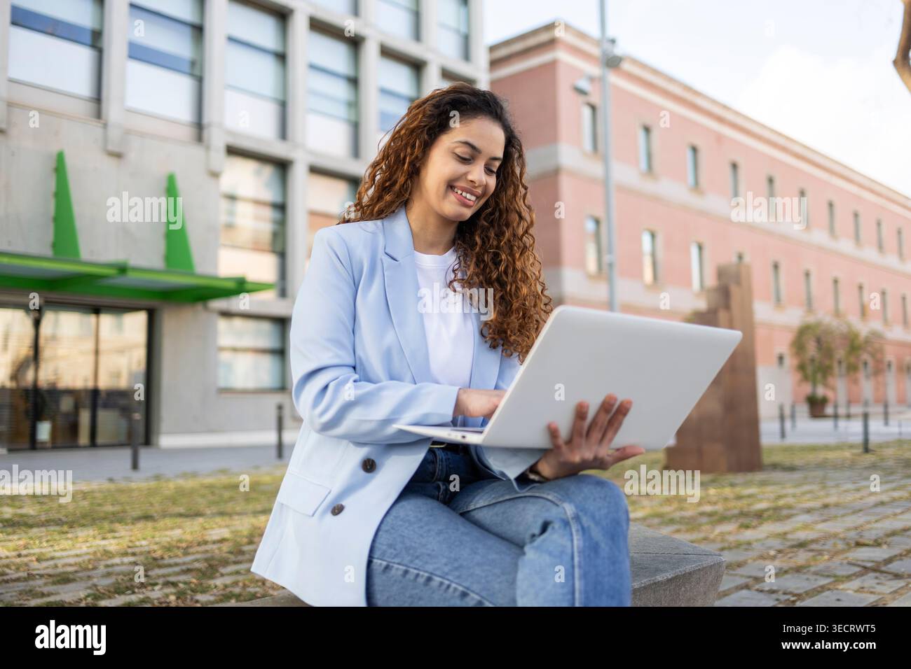 Una donna d'affari sorridente in un blazer leggero lavora su un notebook mentre si siede fuori da un moderno complesso di uffici, mostrando lavoro remoto e comunicazione digitale Foto Stock