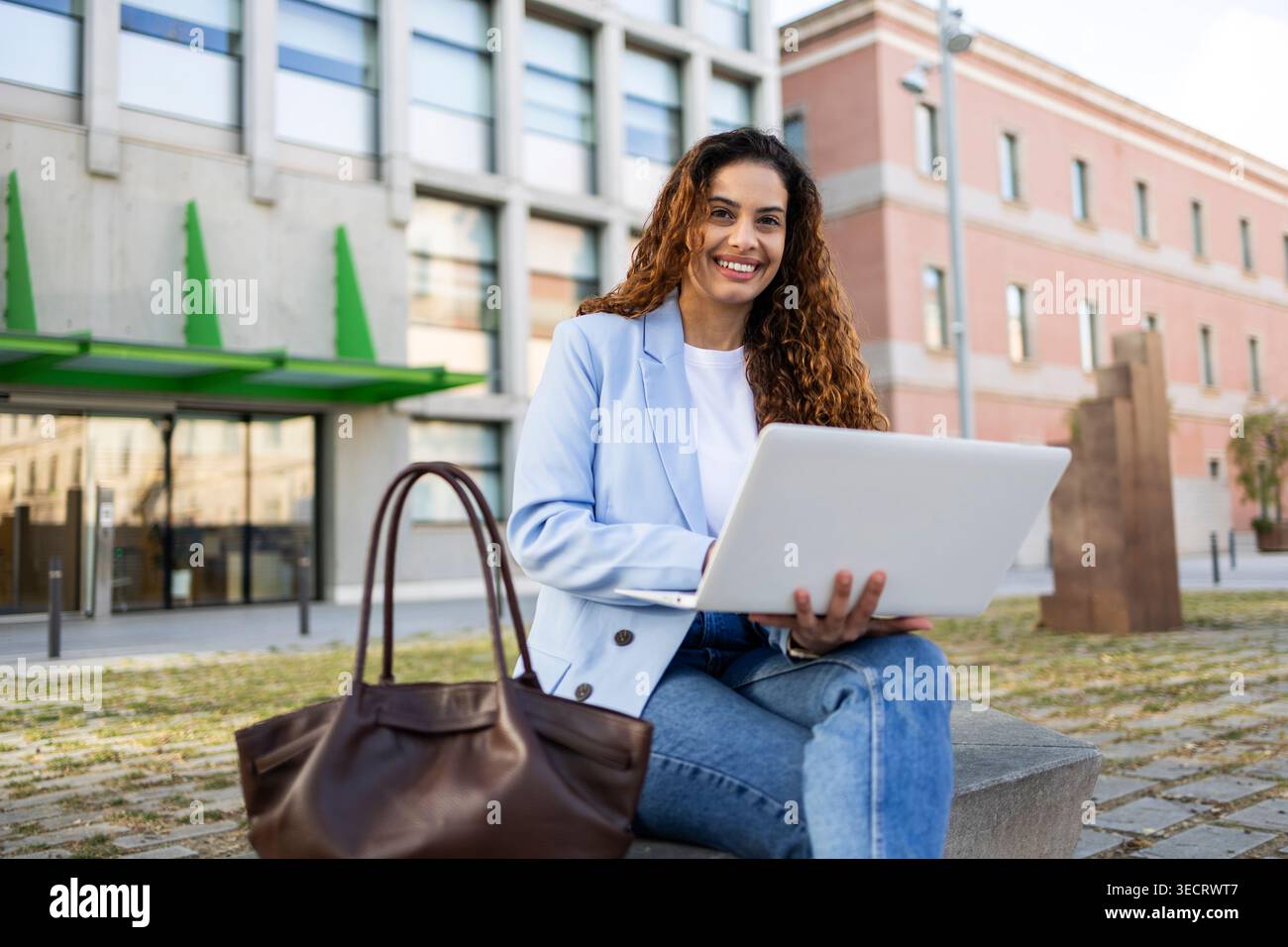Donna d'affari sorridente, seduta all'aperto con un laptop e una borsa in pelle, che lavora a distanza vicino a un moderno edificio di uffici alla luce del giorno, mescolando vita urbana Foto Stock