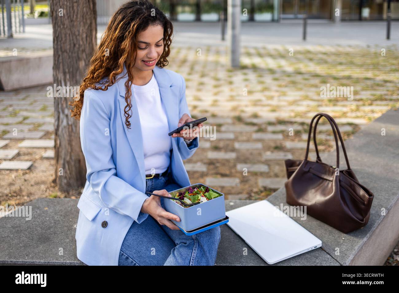 Donna d'affari sorridente in un blazer blu controlla il suo smartphone mentre mangia un sano pranzo con insalata all'aperto, con laptop e borsa nelle vicinanze, bilanciando WO Foto Stock