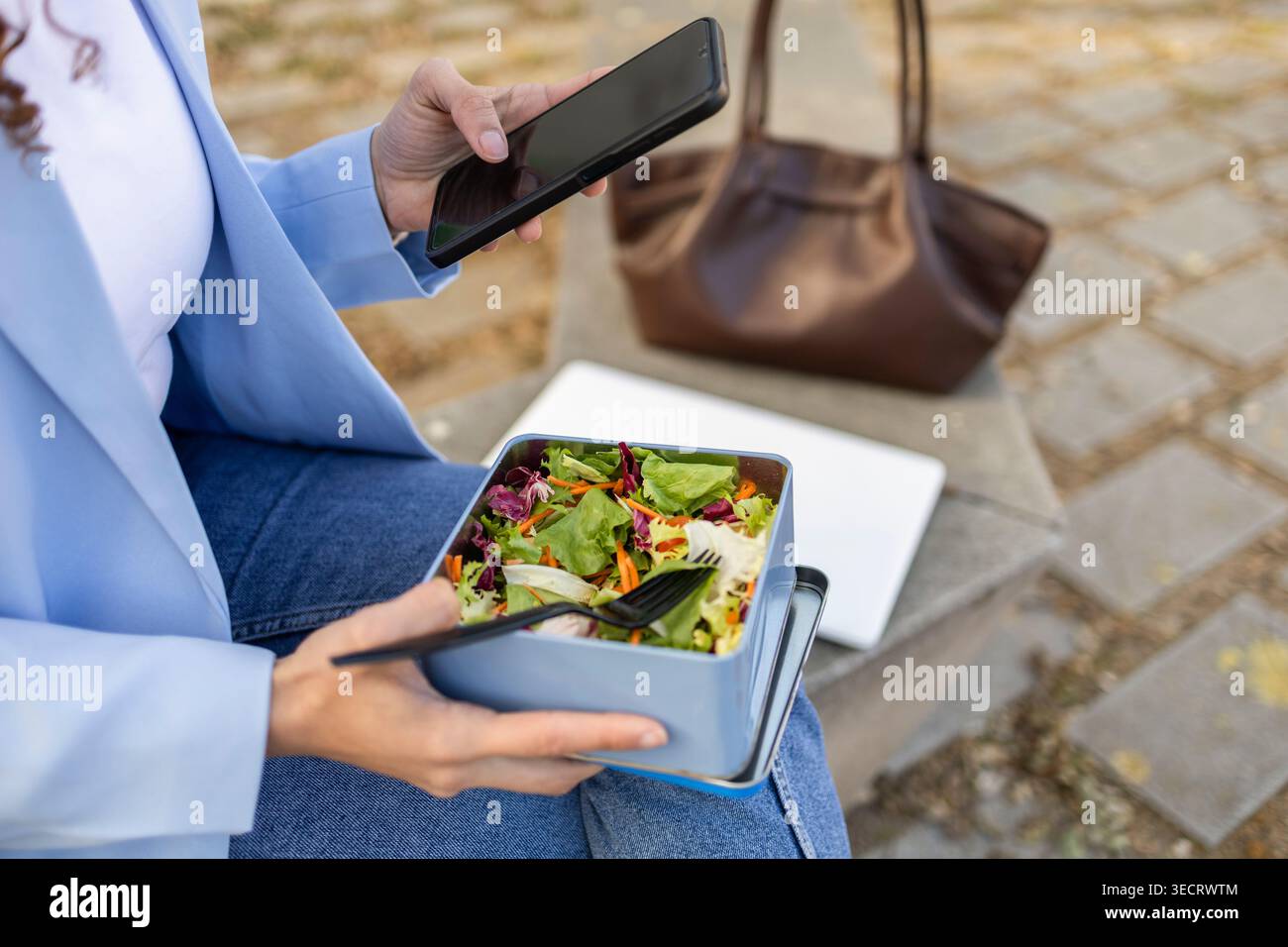 Primo piano di una donna d'affari in un blazer che utilizza uno smartphone mentre mangia un'insalata fresca da un cestino per il pranzo all'aperto, con laptop e borsa nelle vicinanze durante Foto Stock