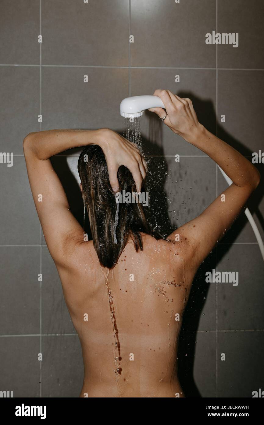 Vista posteriore di una donna che si lava i capelli sotto la doccia, l'acqua che scorre sulla schiena e le mani che massaggiano lo shampoo. Le piastrelle minimali del bagno creano un ambiente pulito Foto Stock